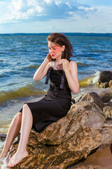 Relaxing Caucasian Brunette Girl Posing in Black Dress On Stone Line At Sea During Sunny Day