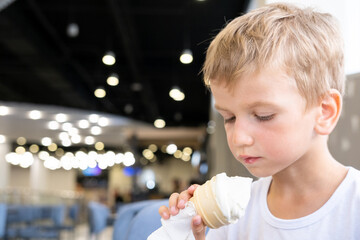 Portrait of a small hungry funny boy eating cold delicious ice cream in a waffle cup sitting in a...