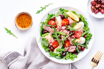 Nicoise salad with tuna, red tomatoes, boiled eggs, green beans and olives on plate, white table background, top view