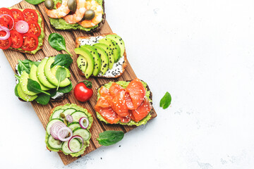 Avocado sandwiches or toasts with salmon, shrimps, tomatoes, cucumbers, soft cheese and spinach, cashew nuts and sesame seeds on wooden cutting board, white table background, top view