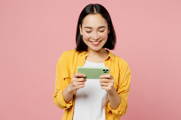 Fototapeta premium Young fun gambling woman of Asian ethnicity wear yellow shirt white t-shirt use play racing app on mobile cell phone hold gadget smartphone for pc video games isolated on plain pastel pink background.