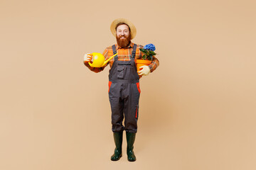 Full body young bearded man wear straw hat overalls work in garden hold blue hydrangea flower in pot and water can isolated on plain pastel beige color background studio portrait Plant caring concept.