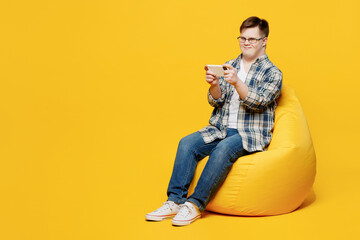 Full body young man with down syndrome wear glasses casual clothes sit in bag chair use play app...