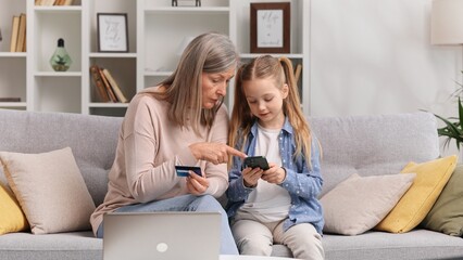 Young granddaughter teaches her senior grandmother to use a card for shopping on the Internet. A 60s grandmother and a little blond granddaughter are buying something by phone payment.