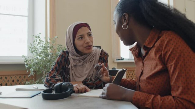 Cheerful young Black and Middle Eastern women sitting at table in classroom doing grammar exercise together