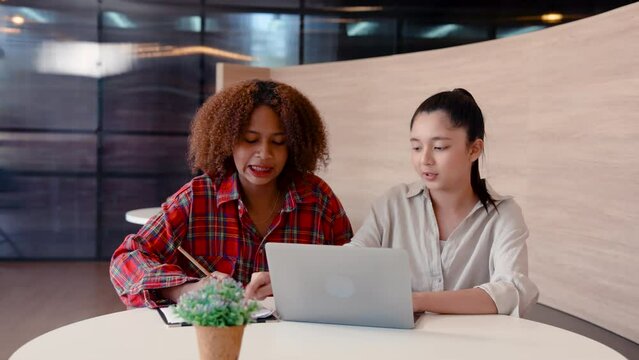 4K, girl African Asian faces who are classmates, sit help each other study exam books before final exam tomorrow, sit study for exam with stern look with eyes staring at laptop computer screen.