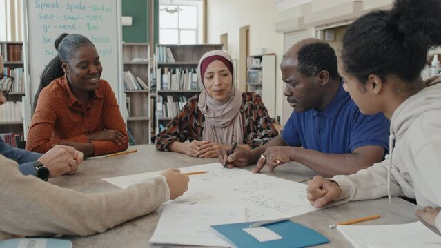 Group of multi-ethnic migrant students sitting at table in library making educational poster during language class