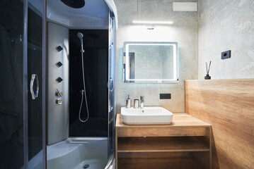 Minimalist grey bathroom with modern shower cabin and white sink on wooden countertop inside. Front view of ceramic basin and mirror with warm back lights turned on in bathroom. Concept of interior.