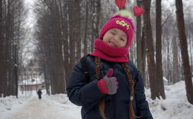 A baby girl in winter warm clothes stands in a winter park and shows her hand likes. Selective focus