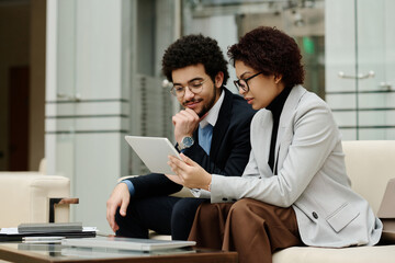 Business colleagues discussing online presentation on tablet pc together while sitting on sofa in lobby during meeting