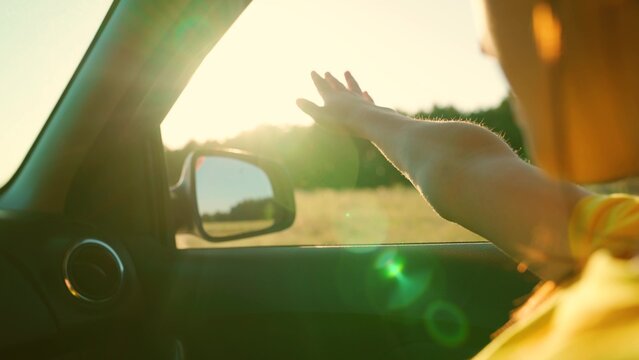 Young woman travels by car catches wind with her hand from car window. Girl with long hair is sitting in front seat of car, stretching her arm out window and catching glare of setting sun. Vacation