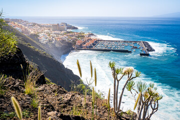 Panoramic view from a high cliff captures the picturesque Los Gigantes village, Puerto de Los Gigantes harbor and Playa de Los Guíos beach with the Atlantic Ocean and island La Gomera in the distance.