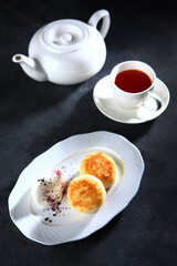 Sweet breakfast - cheese pancakes. Cottage cheese pancakes with sour cream on a white plate. Dark background.A cup of tea.Vertical photo.