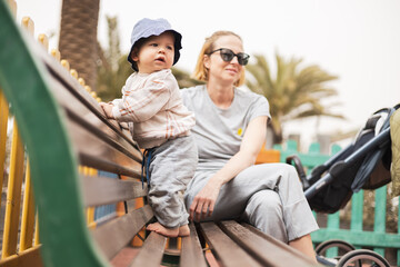 Young mother with her cute infant baby boy child on bench on urban children's playground on warm...