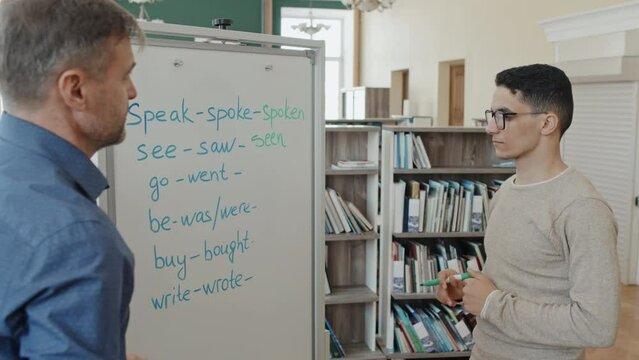 Young Middle Eastern man attending English language class for immigrants writing past participle forms of irregular verbs on whiteboard