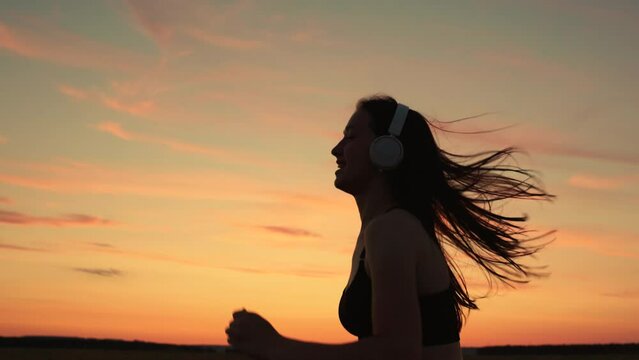 Training run. Free young woman runs in summer in park at sunset, listens to music on headphones. Silhouette, beautiful girl in musical headphones, is engaged in fitness, runs in evening. Teen runner