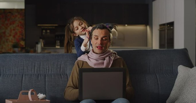Funny Family Situation: Portrait of a Man Having a Video Call Conference with his Colleagues Using Laptop Computer at Home While his Daughter is Playing Makeup on him. Work from Home Concept