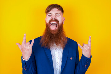 red haired man wearing blue suit over yellow studio background making rock hand gesture and showing...