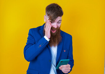red haired man wearing blue suit over yellow studio background holding in hands cell reading sms using new app 5g
