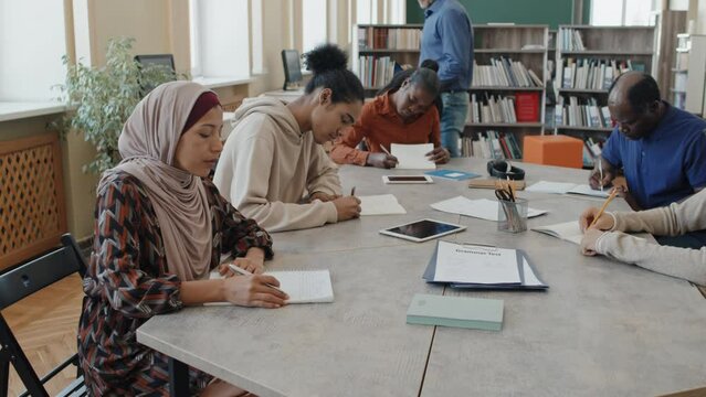 Muslim woman wearing hijab and multi-ethnic groupmates sitting at table working on writing task during language class for migrants