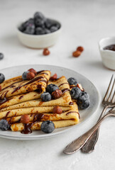 Crepes, blinis or thin pancakes with blueberries, hazelnuts and chocolate sauce on a white plate at white stone table. Maslenitsa traditional festival meal. Delicious breakfast