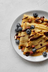 Crepes, blinis or thin pancakes with blueberries, hazelnuts and chocolate sauce on a white plate at...