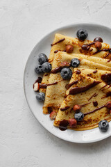 Crepes, blinis or thin pancakes with blueberries, hazelnuts and chocolate sauce on a white plate at white stone table. Maslenitsa traditional festival meal. Delicious breakfast