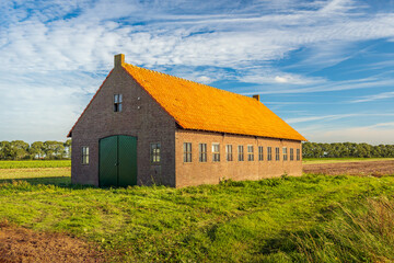 Fototapeta na wymiar Brick barn with an orange tiled roof in the foreground in farmland. The photo was taken in the Dutch province of North Brabant on a sunny day at the beginning of the autumn season.
