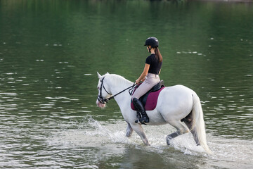 Girl riding a snow white horse down the calm river water with forest greenery reflections from the...