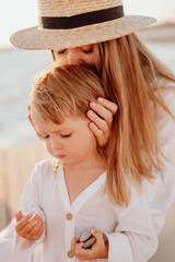 A beautiful young girl with long hair in a white dress and a straw hat kisses her little daughter's head on the background of the evening ocean