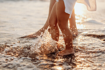 The bare feet of an adult woman and a girl, a child in white dresses are splashing in the ocean at...