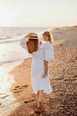 A small child, a girl in her mother's arms, both in white dresses, walk barefoot along the ocean at sunset.