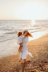 A small child, a girl in her mother's arms, both in white dresses, walk barefoot along the ocean at sunset.
