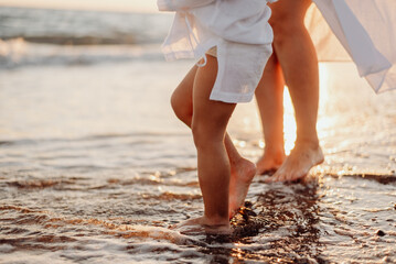 The bare feet of an adult woman and a girl, a child in white dresses stand in the water on the ocean at sunset. Water drops sparkle in the sun