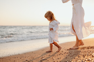 A little girl and a young mother in white dresses and a hat are walking on the water on the ocean at sunset. Deserted beach.