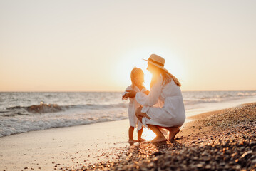 A young mother sat down and holds her little daughter by the hand on the ocean at sunset. Girls in white dresses and long hair.