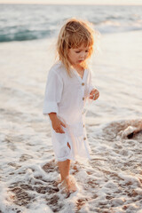 A little girl with long hair, in a white dress, walks barefoot along the ocean. The feet are washed by the sea with foam. Blurred ocean background at sunset.