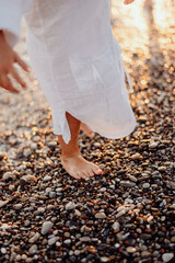 A little girl with long hair, in a white dress, walks barefoot along the ocean. Feet stand on the shore with round small stones.