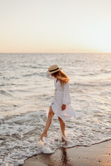 A young, beautiful girl with long hair in a straw hat and a white dress dances on the water near the sea at sunset. Blurred background.