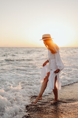 A young, beautiful girl with long hair in a straw hat and a white dress stands in the water on the ocean at sunset. Blurred background. wave crashes on the shore