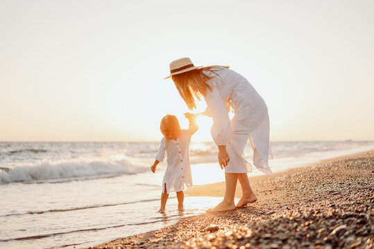 A young mother holds her little daughter by the hand and together they walk along the ocean towards the sunset. Girls in white dresses and with long hair that develops the wind