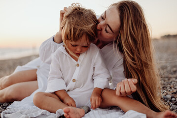 A young mother and daughter in white dresses are sitting on a rocky ocean shore at sunset. A little daughter looks at a stone in her hands, and her mother kisses her