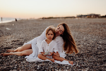 A young mother and daughter in white dresses are sitting on a rocky ocean shore at sunset. Little daughter looks ahead and her mother kisses her