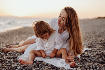 A young mother and daughter in white dresses are sitting on a rocky ocean shore at sunset. Little daughter looks at the stone in her hands