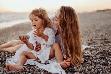 A young mother and daughter in white dresses are sitting on a rocky ocean shore at sunset. Little daughter looks at the stone in her hands