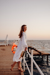 A young girl with long hair in a white dress stands on a wooden pier and holds on to a white handrail against the backdrop of the ocean at sunset. Hat in hand. Hair develops the wind