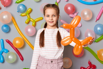 Smiling child. Birthday party. Delighted satisfied little girl with braids standing against gray...