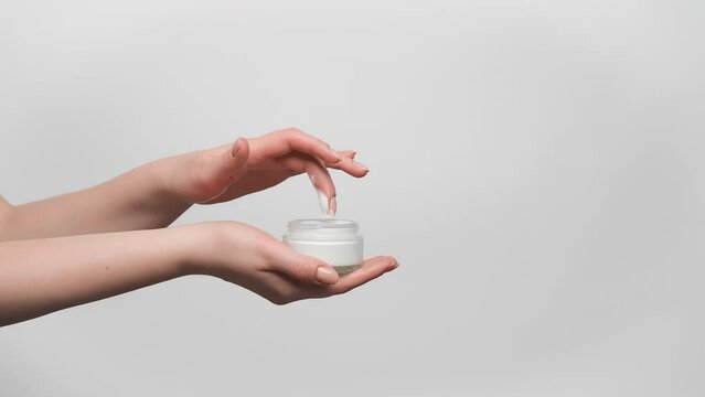 Woman is holding glass jar of face cream in her hand, taking a sample with a finger, on gray background, copy space. 4k, slow motion.