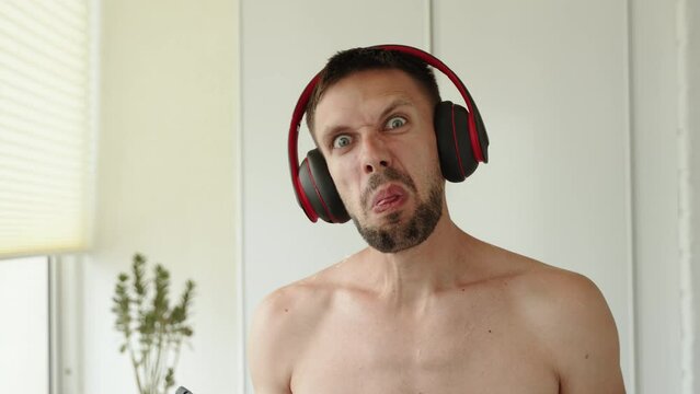 Portrait of naked man in wireless headphones stands at home and teases, shows tongue at camera. Thin guy with dark beard shakes phone in displeasure, makes nasty faces, boasts modern equipment.