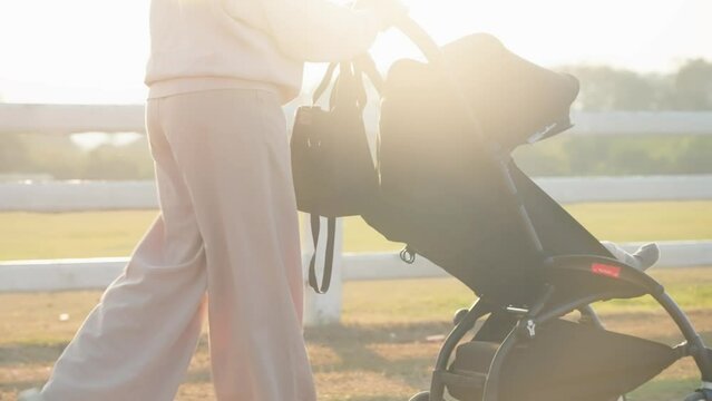 Shot of mother pushing baby stroller car walking around the public area during sunset with beautiful sunlight leak, warm family vibe, new mother and how to raise a child, modern gear for convenience